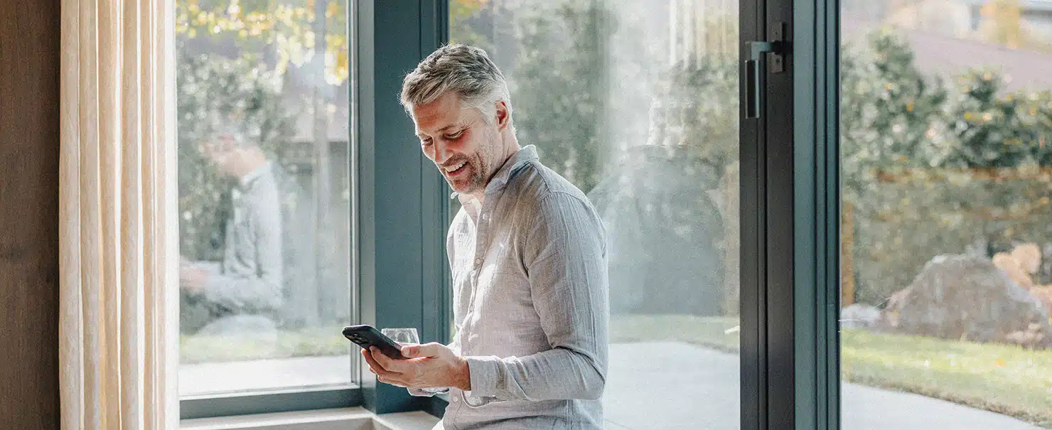 Ein lächelnder Mann steuert im modernen Wohnzimmer den KATZBECK Sonnenschutz bequem über sein Smartphone via Smart Home App.