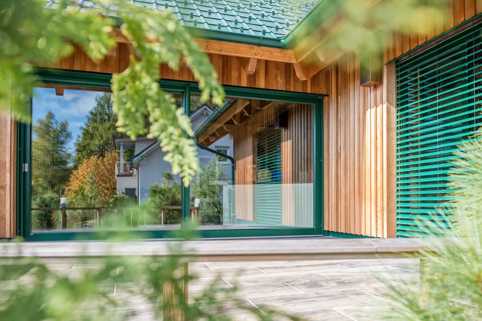 Nahaufnahme der Fassade eines modernen Holzhauses mit KATZBECK Holz-Alu-Fenstern in einer harmonischen grünen Außenfarbe und dazu passenden grünen Raffstores, die sich in die Natur einfügen.